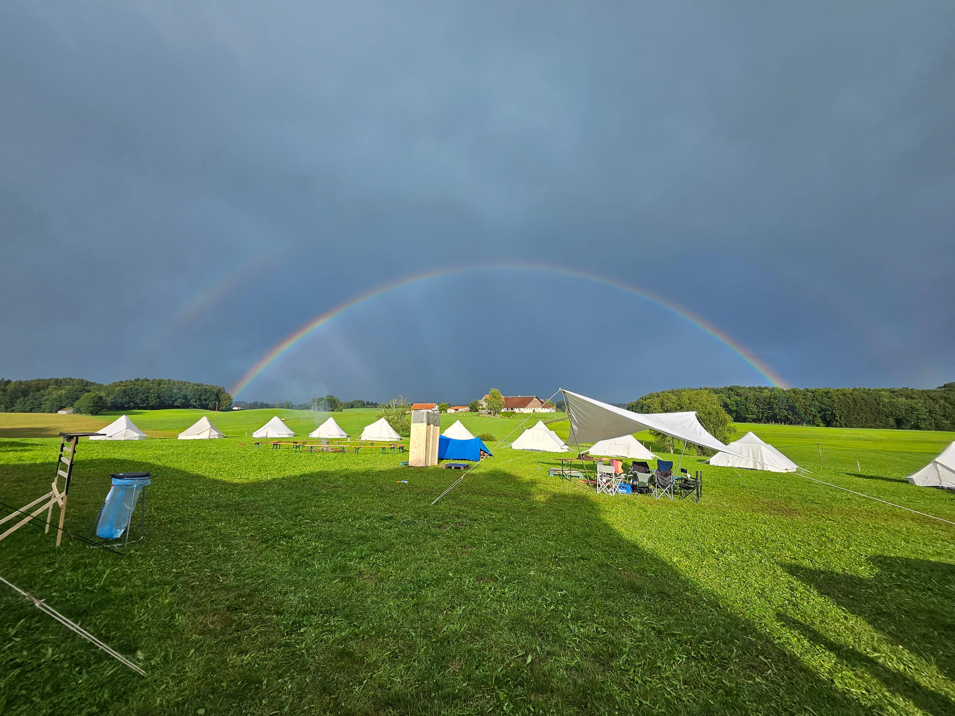 Zeltplatz des Boni-Arche-Camps unter einem Regenbogen nach einem Gewitter, mit weißen Zelten auf grüner Wiese.