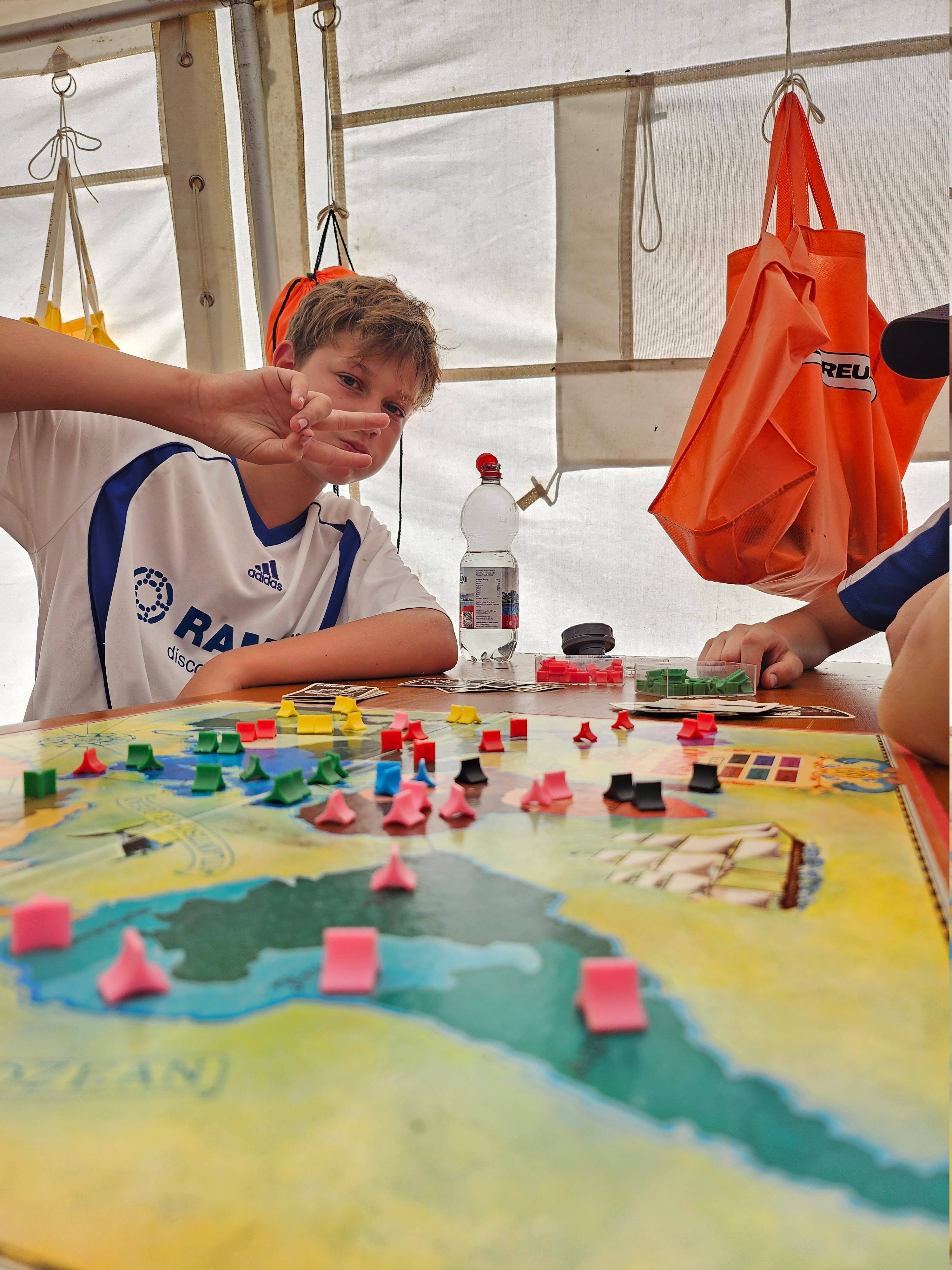 Junge spielt im Zelt ein buntes Brettspiel mit farbigen Figuren und macht eine spielerische Handgeste.
