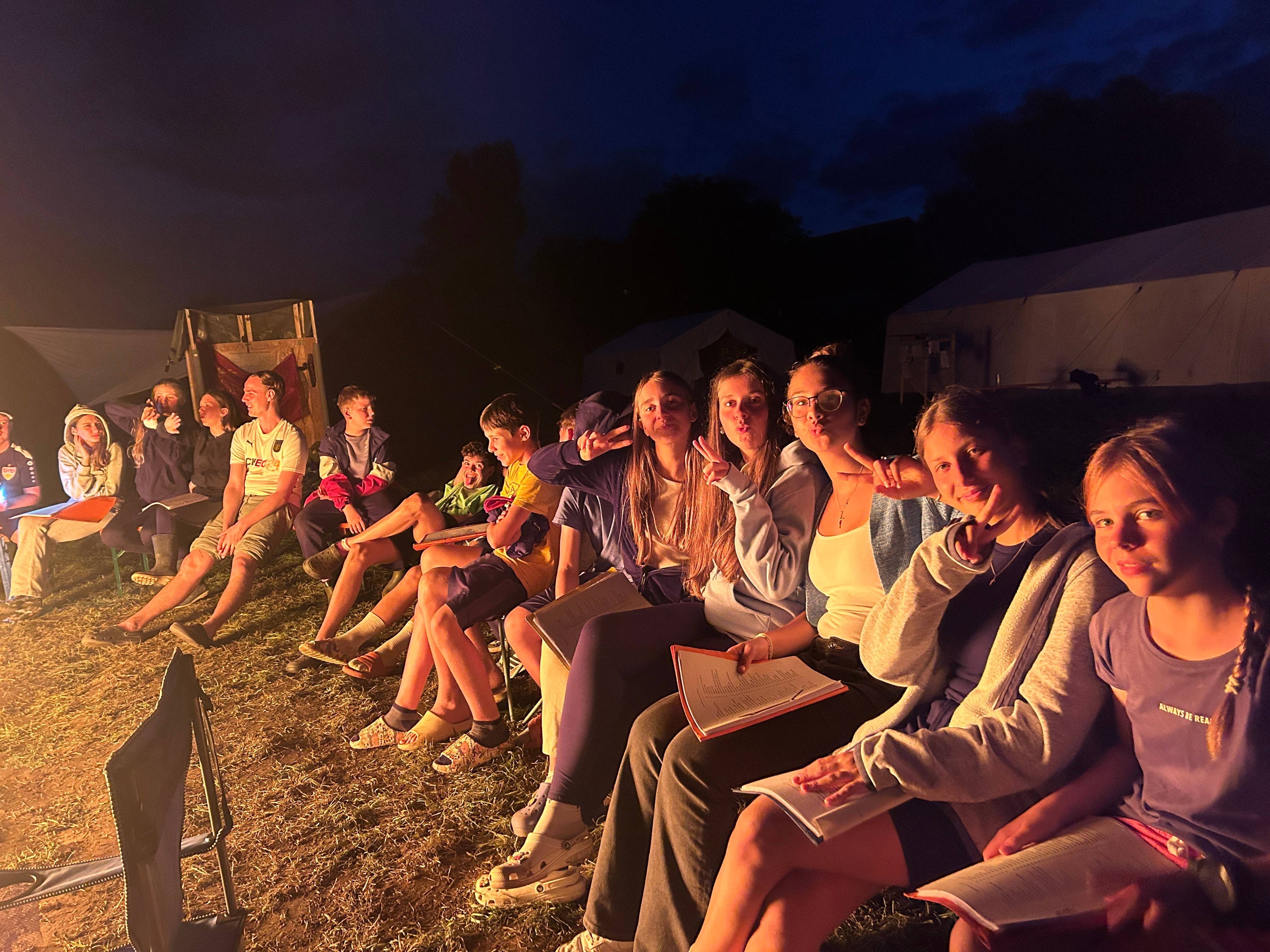 Jugendliche beim Abendprogramm im Zeltlager, mit Notizblöcken und Grimassen vor dem dunklen Himmel.