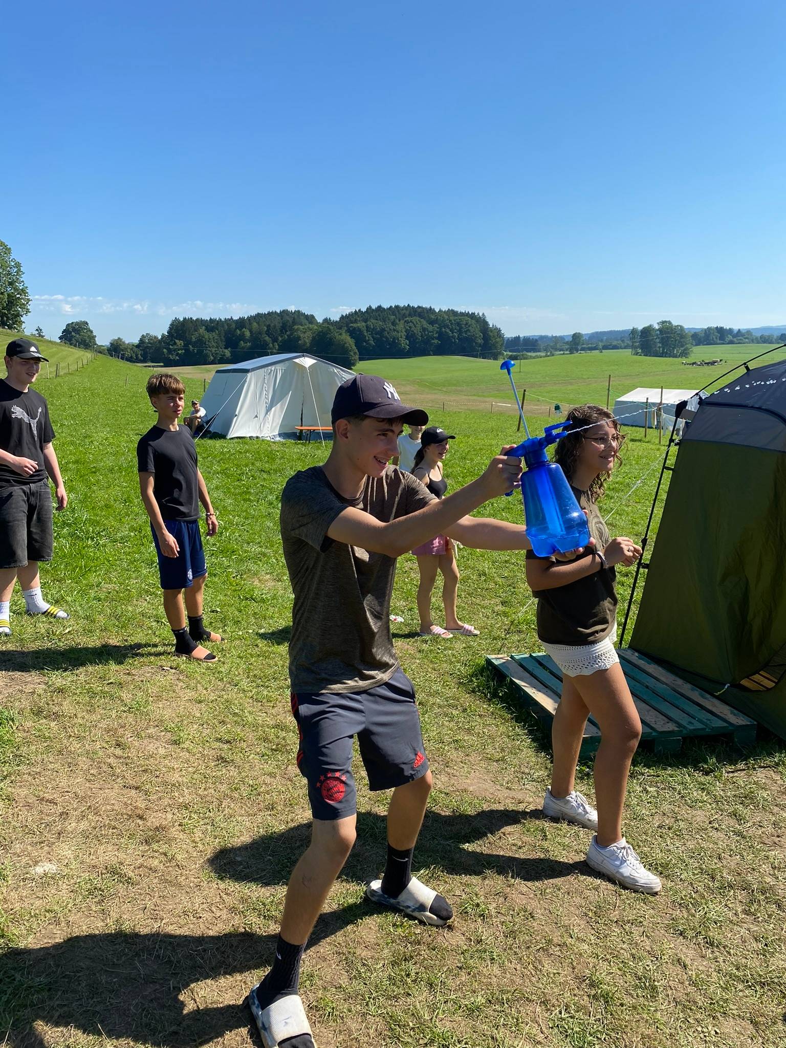 Jugendliche spielen mit einer Wassersprühflasche auf der Wiese des Boni-Arche-Camps, im Hintergrund Zelte und grüne Hügel.