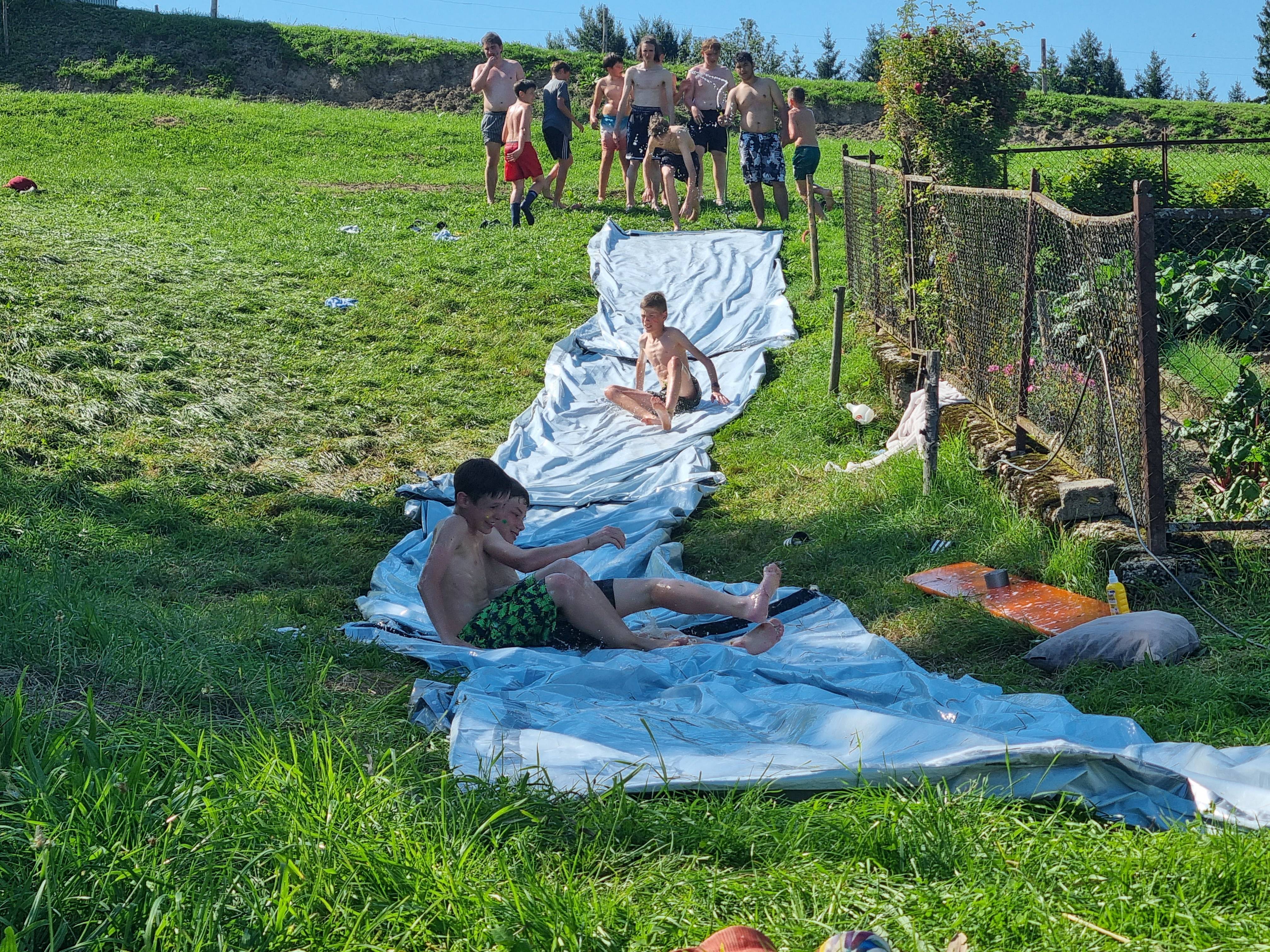 Kinder rutschen lachend auf einer nassen Folie bergab während eines Wasserspiels im Boni-Arche-Camp.