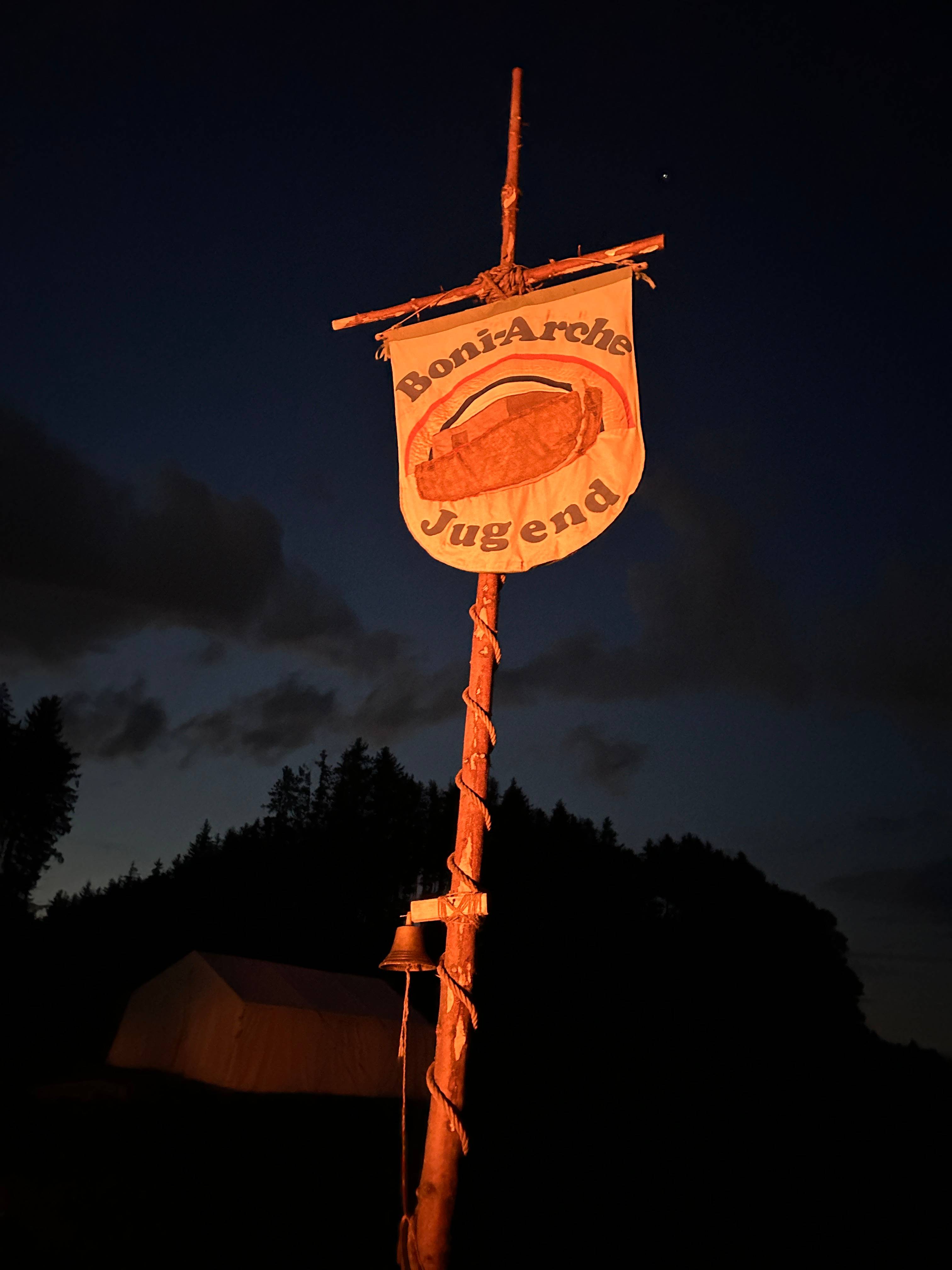 Das beleuchtete Banner der Boni-Arche Jugend weht nachts am Lagermast, im Hintergrund das Zelt und die dunkle Waldsilhouette.