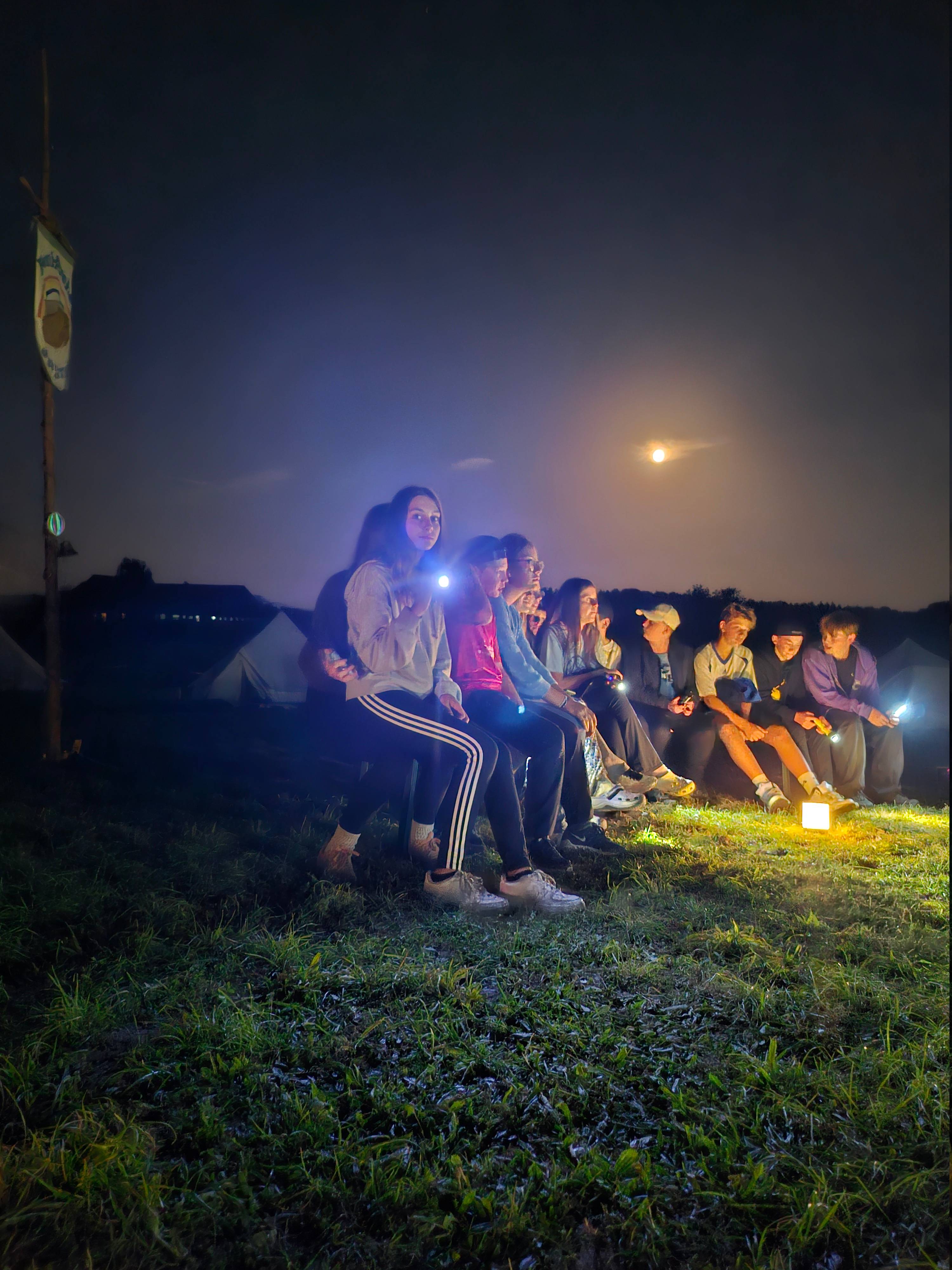 Jugendliche sitzen nachts im Gras, leuchten mit Taschenlampen und genießen das Lagerprogramm unter dem Mond.