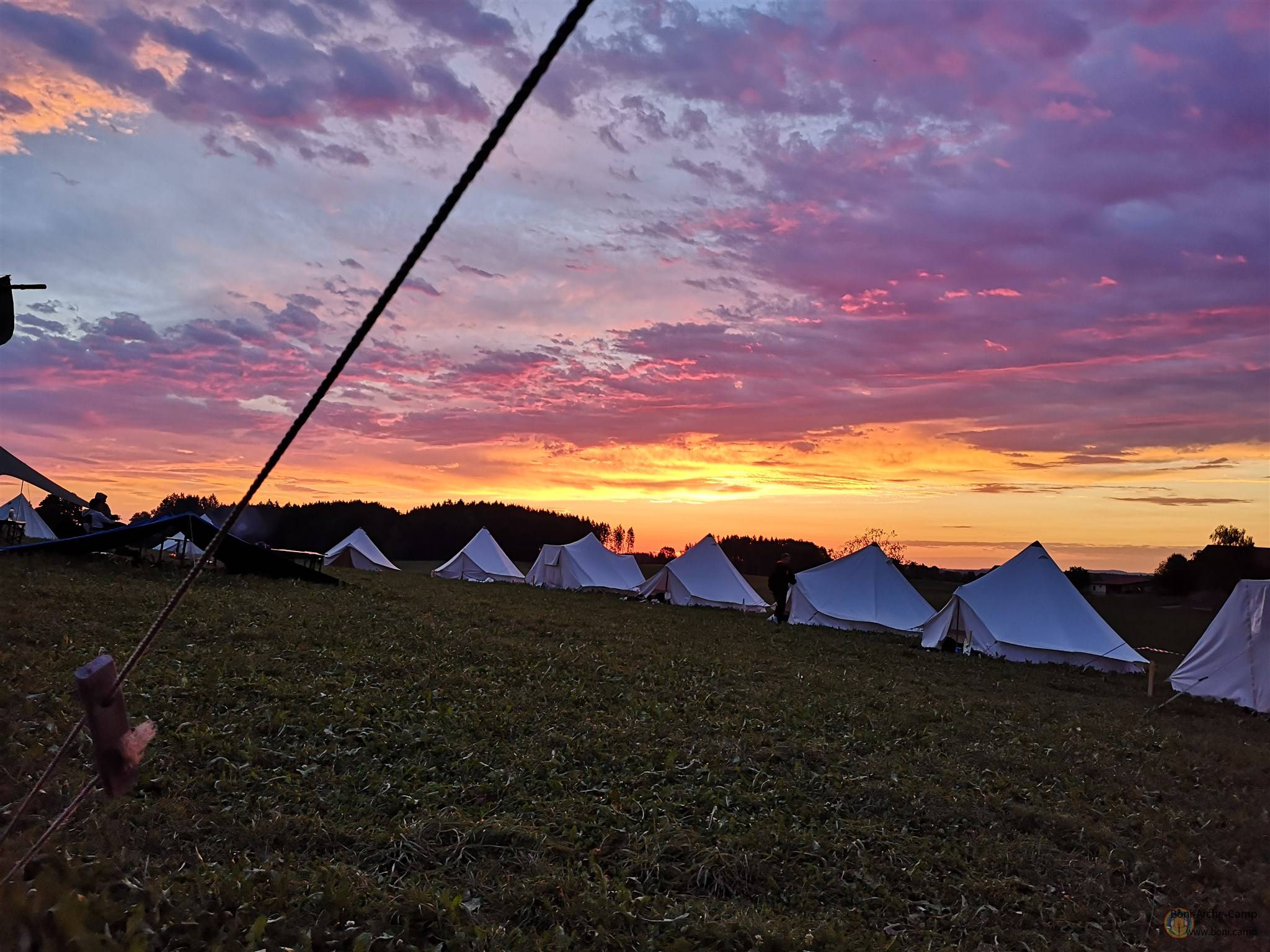 Zeltplatz des Boni-Arche-Camps bei Sonnenuntergang mit farbenfrohem Himmel und weißen Zelten auf der Wiese.