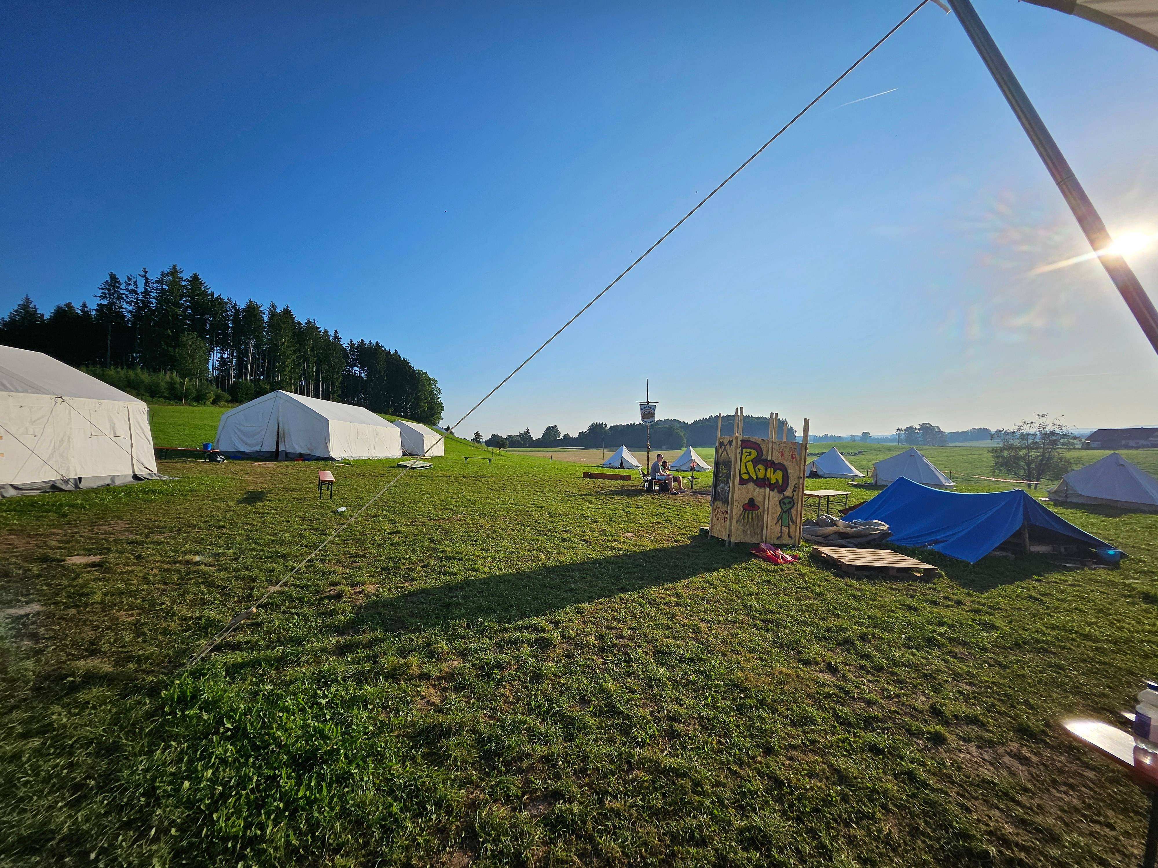 Boni-Arche-Camp: Zeltplatz mit weißen Gruppenzelten, blauem Zelt und selbstgebautem 'Rom'-Schild auf einer sonnigen Wiese.