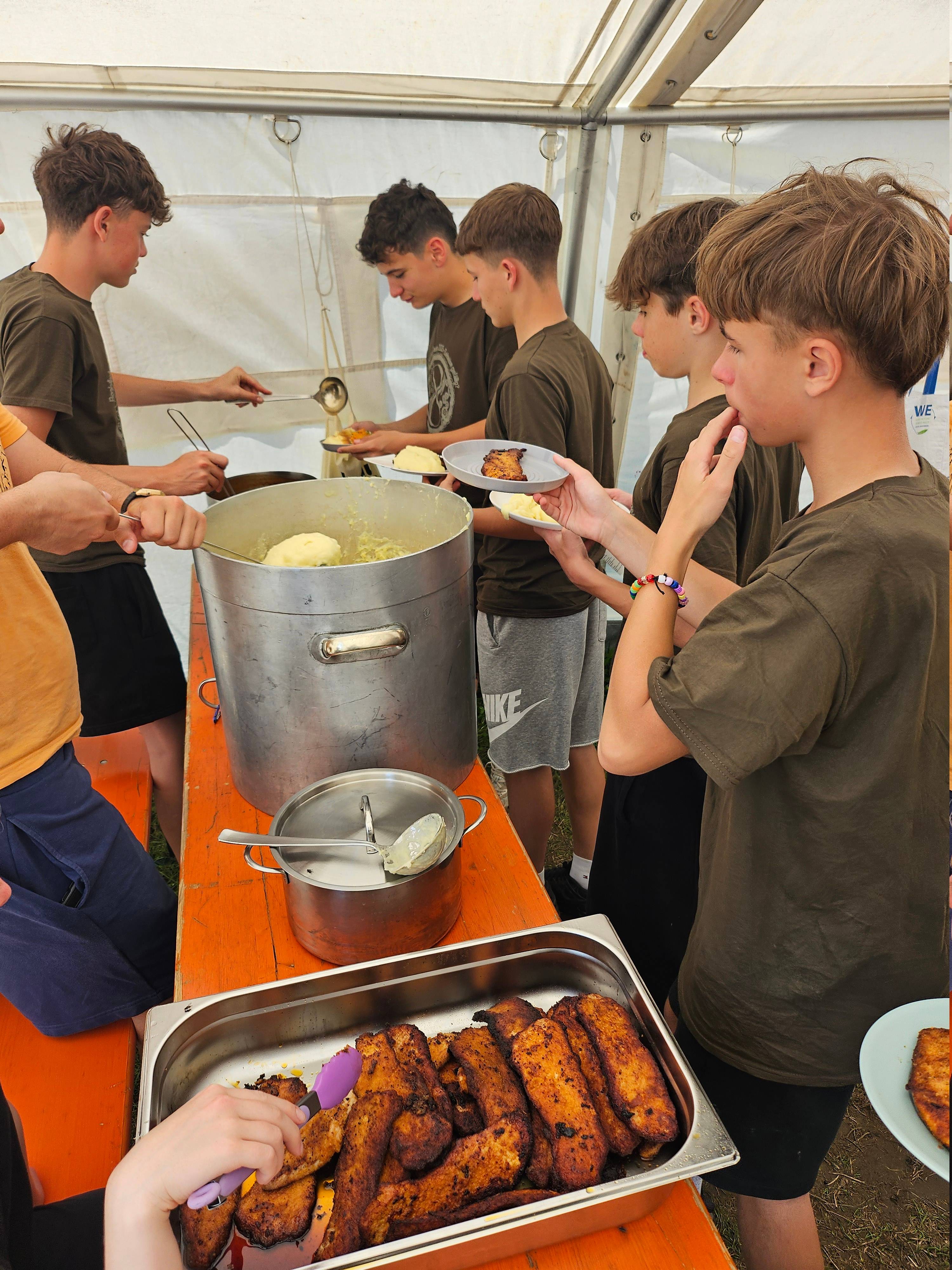 Jugendliche holen sich Essen aus großen Kesseln und Wannen an einem Tisch unter einem Zelt beim Boni-Arche-Camp.