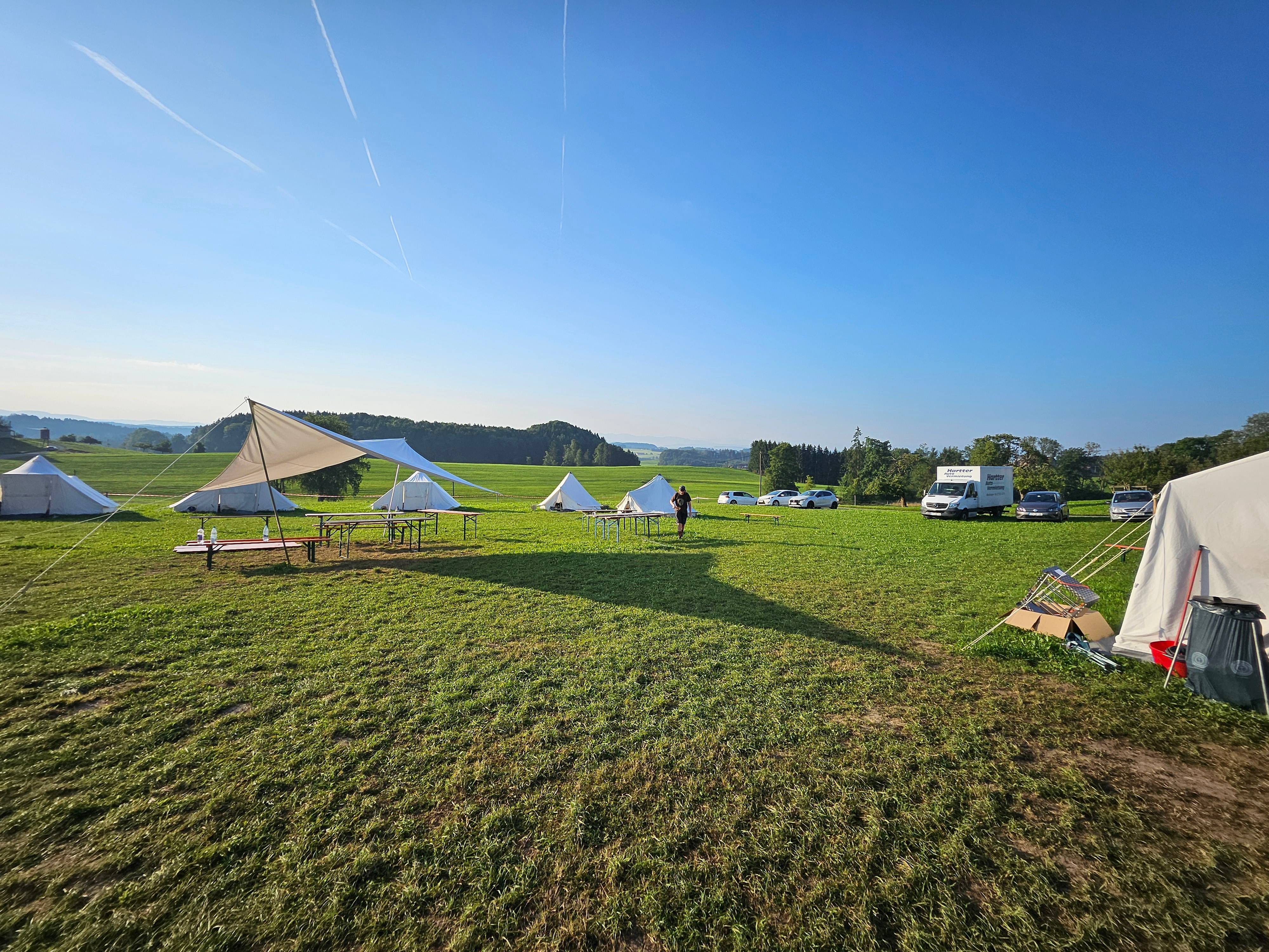 Weitläufiger Zeltplatz des Boni-Arche-Camps mit weißen Zelten, Sonnensegel und Tischen auf einer grünen Wiese unter blauem Himmel.