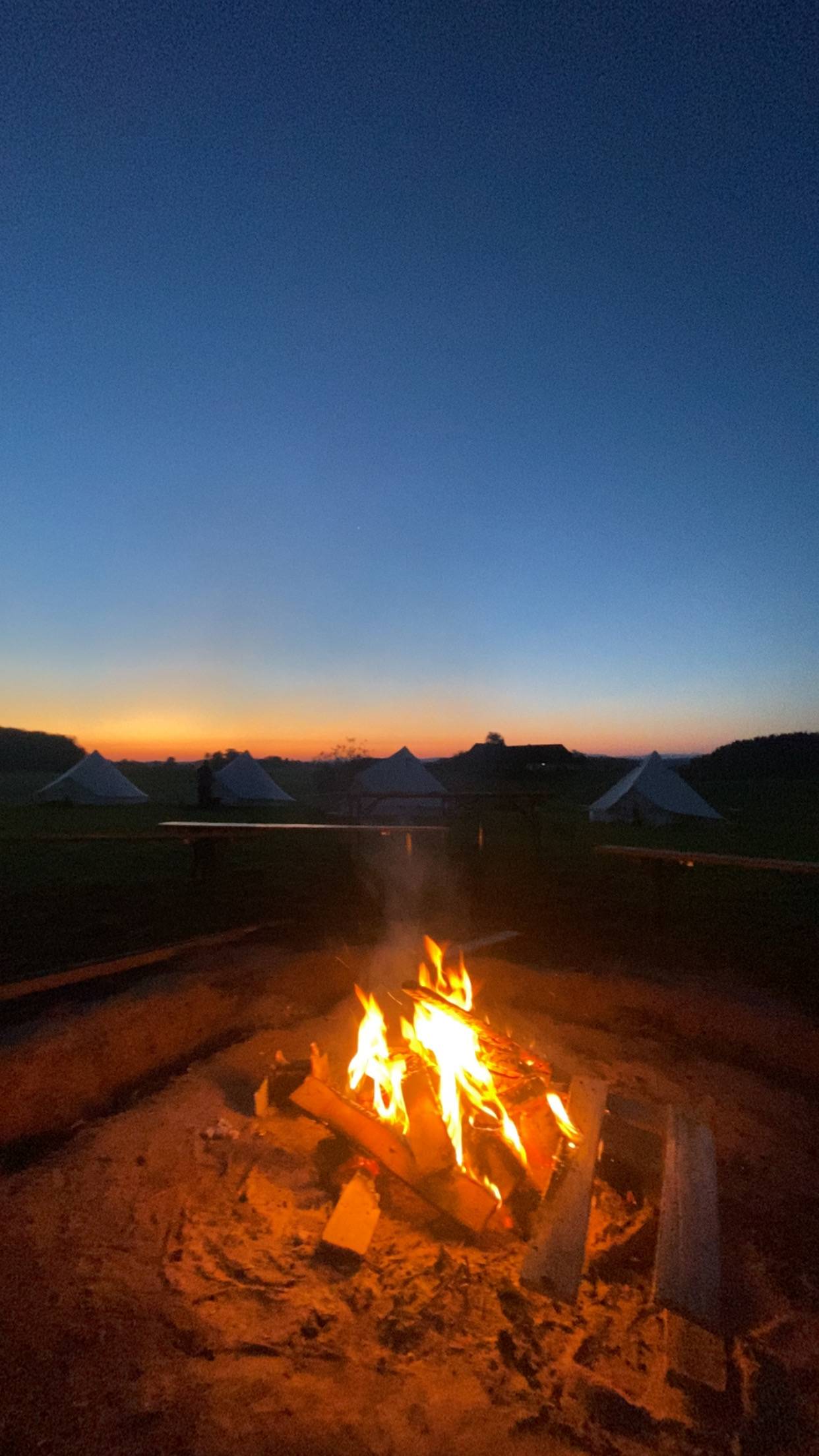 Lagerfeuer leuchtet im Abenddämmerung vor den Zelten des Boni-Arche-Camps, während der Himmel in warmen Farben erstrahlt.